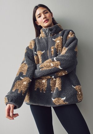 Femeie care poartă o geacă gri oversized din fleece cu modele de tauri maro și albi, stând în fața unui perete simplu gri deschis.
