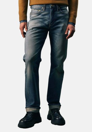 Mann trägt ausgewaschene blaue Jeans mit umgekrempelten Säumen, schwarze klobige Schuhe, einen braunen Pullover und ein blaues Hemd darunter, steht vor einem schlichten Hintergrund.