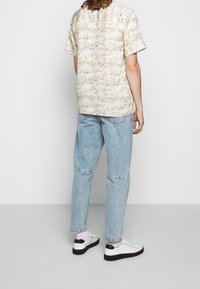 Camicia a maniche corte beige con motivo, jeans in denim azzurro chiaro e sneakers bianche con dettagli neri. Outfit casual con vestibilità rilassata.