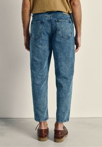 Svetlo modri traper hlače z ohlapnim krojem, z zadnjimi žepi, rahlo zoženimi hlačnicami in raztrganimi robovi. Nosi se z rjavimi klompi.