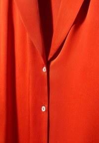 Blusa rossa in un tessuto leggero e morbido. Presenta un colletto, una chiusura con bottoni bianchi e una vestibilità leggermente ampia.