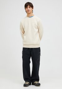 Felpa beige con scollatura rotonda e polsini a costine, abbinata a pantaloni cargo neri larghi con tasche con zip.