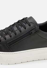 Baskets en cuir noir avec lacets noirs et fermeture éclair latérale, contrastées par une semelle en caoutchouc épaisse blanche.