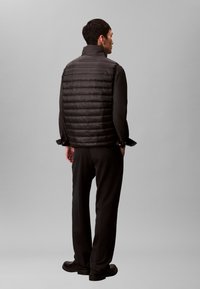 Čierna prešívaná vesta s vysokým golierom, vyznačujúca sa elegantným dizajnom a jemným leskom. Nosená cez tmavú dlhú košeľu a nohavice.