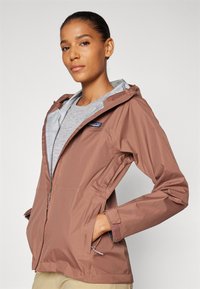 Chaqueta cortavientos marrón con capucha, que cuenta con un forro gris, bolsillos con cremallera y puños ajustables. Tejido ligero con un diseño elegante.