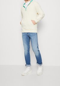 Sweat à capuche crème à fermeture éclair avec des accents bleu sarcelle, poches avant, associé à un jean bleu clair et des baskets blanches. Coupe décontractée, texture douce.