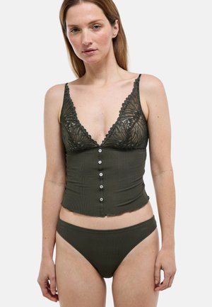 Femme portant un ensemble de lingerie vert foncé avec un top camisole bordé de dentelle et un slip assorti, debout sur un fond clair.