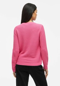 Roze gebreide trui met een gestructureerd honingraatpatroon, geribbelde manchetten en zoom, gecombineerd met zwarte broek. Gezicht vanuit de achterkant.