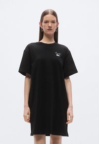 Čierne bavlnené tričko-šaty s krátkymi rukávmi, okrúhlym výstrihom a malým bielym logom na ľavej strane hrudníka. Bežný strih, hladká textúra.