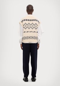 Pull sans manches en maille crème avec des motifs géométriques bleu marine, porté sur une chemise blanche à manches longues, assorti à un pantalon noir ample.