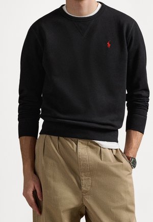 Man draagt zwarte trui met rood logo op de borst, een wit ondershirt ingestopt en beige geplooide broek, met één hand in de zak.