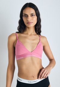 Ružova čipkasta braletka z V-izrezom in tankimi trakovi. Ima blagovno znamko obroč z napisom "Calvin Klein." Gladka tekstura, oprijeta oblika.