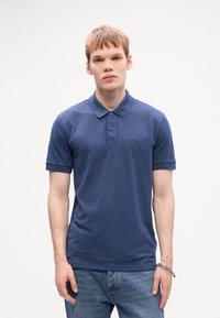 Joven vistiendo una camiseta polo de manga corta azul con el logo "BOSS" y jeans azules, de pie contra un fondo claro y liso.