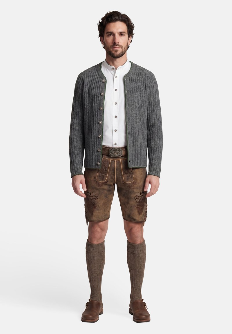 Mann in traditioneller bayerischer Tracht: grauer, geknöpfter Cardigan, weißes Hemd, braune, bestickte Lederhose, knielange Socken und braune Schuhe.