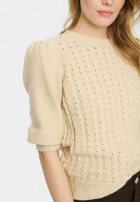 Bežs adīts džemperis ar īsām puff piedurknēm, teksturētu dimanta rakstu un ribotu apakšmalu. Mīksta auduma ar atvērta adīkšana. 