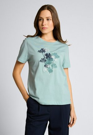 Vrouw met bruin haar die een lichtblauw T-shirt draagt met een donkerblauw en wit bloemenpatroon, gecombineerd met donkerblauwe broek, staand met één hand in de zak.