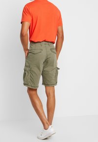 Oliivinvihreät cargo-shortsit sivutaskuilla, suoralla leikkauksella ja näkyvillä ompeluilla, yhdistettynä kirkkaan oranssiin t-paitaan ja valkoisiin lenkkareihin.