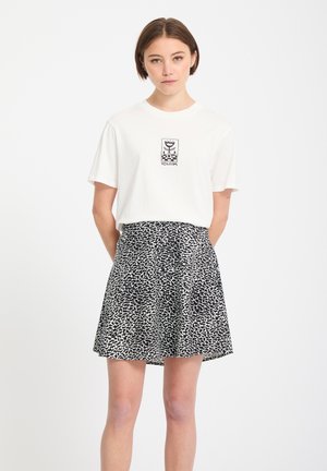 Joven con el cabello corto, vistiendo una camiseta gráfica blanca y una falda con patrón en blanco y negro, de pie contra un fondo liso.