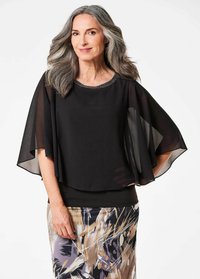 Schwarze Bluse mit durchsichtigen Umhangärmel, perlenverzierter Ausschnitt und geschichteter Säume. Kombiniert mit einem gemusterten Rock in erdigen Tönen.