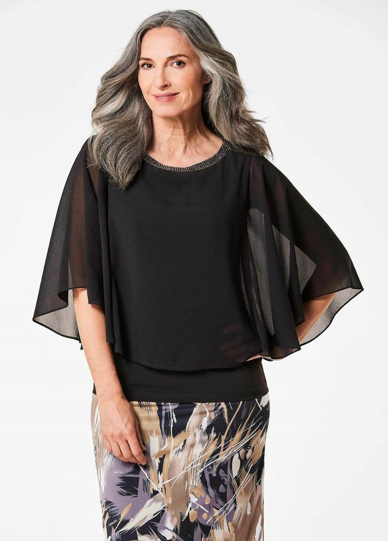 Schwarze Bluse mit durchsichtigen Umhangärmel, perlenverzierter Ausschnitt und geschichteter Säume. Kombiniert mit einem gemusterten Rock in erdigen Tönen.