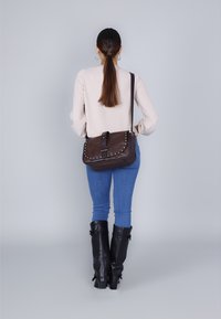 Bruine leren schoudertas met een klep, studs en een band, gedragen over een lichtkleurige top en blauwe jeans. Platte zwarte laarzen zichtbaar.