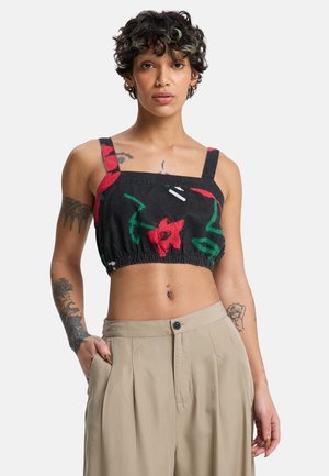 Young person with short curly hair wearing a black floral cropped top and beige wide-leg pants, showing tattoos on arms and torso.