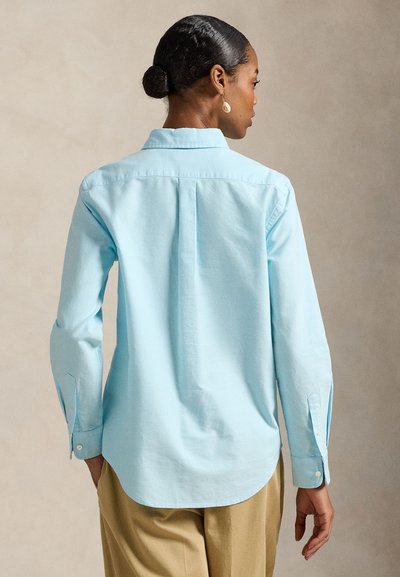 Frau mit dunklen Haaren im Dutt, trägt ein hellblaues Langarmshirt und beige Hosen, von hinten vor neutralem Hintergrund gezeigt.