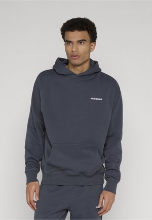 Ung mann iført mørkeblå hettejakke og matchende bukser med små hvite "PEGADOR"-logoer, stående mot en ensfarget lys bakgrunn.