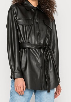 Femme portant une veste-chemise noire en similicuir ceinturée, à manches longues, avec poches poitrine boutonnées et jean bleu.