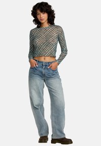 Gjennomsiktig, langermet grønn og blå mønstrete crop top med bølget kant, kombinert med løse lyseblå jeans og brune sko.