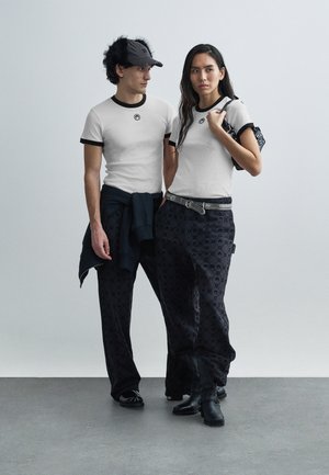 White fitted t-shirts with black trim, paired with black patterned wide-leg pants. One wears a navy sweatshirt, and both have black footwear.