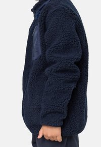 Enfant portant une veste en polaire bleu marine avec un tissu texturé et une poche poitrine zippée, vu de côté avec la main visible.