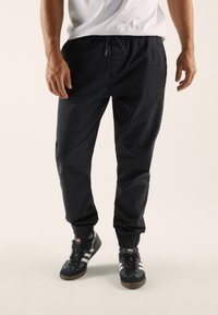 Black, tapered jogger pants with elastic cuffs, drawstring waistband, and smooth fabric, paired with black and white athletic shoes.