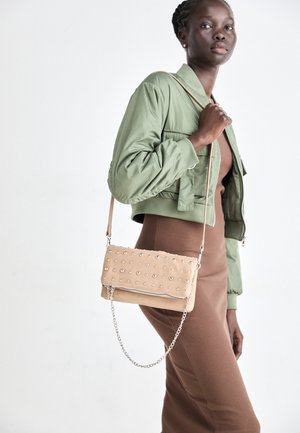 Vrouw draagt een groene cropped jas en een bruine jurk en draagt een beige gestikte handtas met kettingdetail op een witte achtergrond.