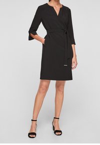 Robe noire avec des manches 3/4, décolleté en V, taille cintrée avec une ceinture, poches latérales, longueur jusqu'aux genoux, en tissu lisse. Assortie avec des sandales à talons noires.