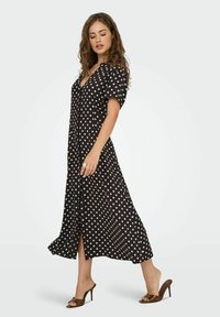 Robe noire jusqu'aux genoux avec des pois blancs, décolleté en V, manches bouffantes et fente latérale. Associée à des sandales à talons hauts marron.