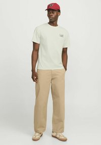 Maglietta verde chiaro con dettagli stampati, pantaloni beige a gamba larga e cappellino da baseball rosso. Le scarpe da ginnastica presentano un accento color sabbia e una base crema.