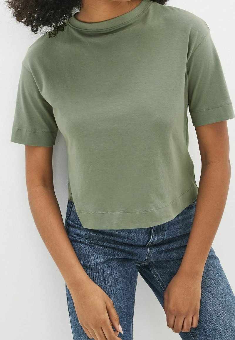 Femme portant un t-shirt court uni vert olive à manches courtes, associé à un jean en denim bleu, debout devant un fond blanc.