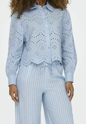 Woman wearing a light blue eyelet button-up blouse with scalloped hem and blue-white striped drawstring pants visible from neck to hips.