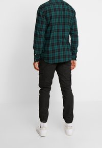 Grün und schwarz kariertes Hemd, lange Ärmel, abgerundeter Saum; kombiniert mit schwarzen Cargo-Hosen und weißen Sneakers mit strukturierten Sohlen.