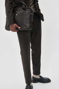 Schwarze Leder-Umhängetasche, die von einer Person in einem dunklen Nadelstreifenanzug gehalten wird. Glatte Textur, rechteckige Form, mit einem Reißverschluss und oberen Griffen.