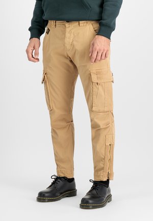Mand iført beige cargobukser med flere lommer og lynlåse, kombineret med sorte snørebåndssko og en mørkegrøn sweatshirt.