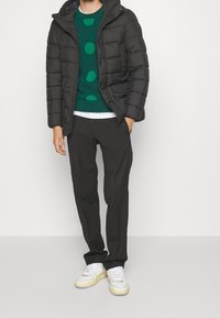 Sort dunjakke, grøn sweater med store prikker, sorte bukser og sneakers med hvid sål og gule detaljer. Casual lag-på-lag outfit.