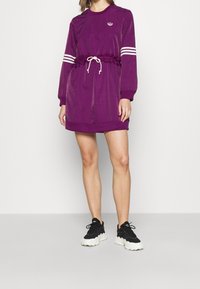 Lila idrottsklänning med långa ärmar, elastisk midja, vit dragsko och tre vita ränder på ärmarna. Svarta sneakers med vita sulor.