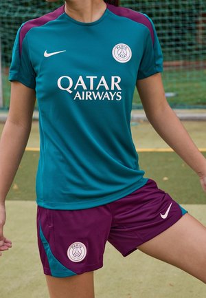 Osoba ubrana w turkusowo-fioletowy strój treningowy Paris Saint-Germain z logo Qatar Airways na boisku z siatką w tle.