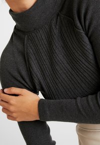 Personne portant un pull à col roulé gris foncé côtelé à manches longues, tenant son bras gauche avec sa main droite sur un fond clair.