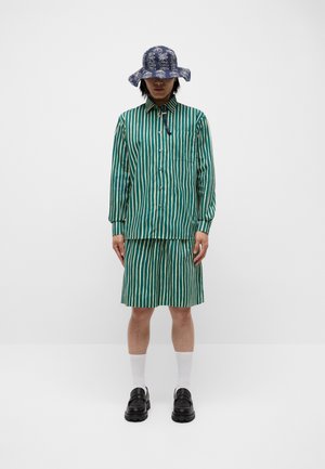 Grön och krämrandig skjorta och matchande shorts, ihop med en marinblå bucket hat med ett mönster. Chunkiga svarta skor och vita strumpor.