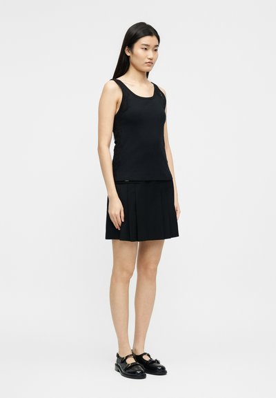 Νεαρή γυναίκα στέκεται, φοράει μαύρο αμάνικο τοπ με λεπτομέρειες από δαντέλα, μαύρη πλισέ φούστα και μαύρα παπούτσια τύπου Mary Jane, μπροστά σε λευκό φόντο.