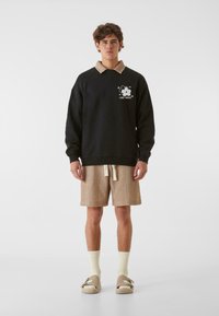 Melna sporta krekls ar beigu apkakli, grafisks ziedu dizains, beigu šorti, gaiši dzelteni zeķes un beigu slip-on apavi.