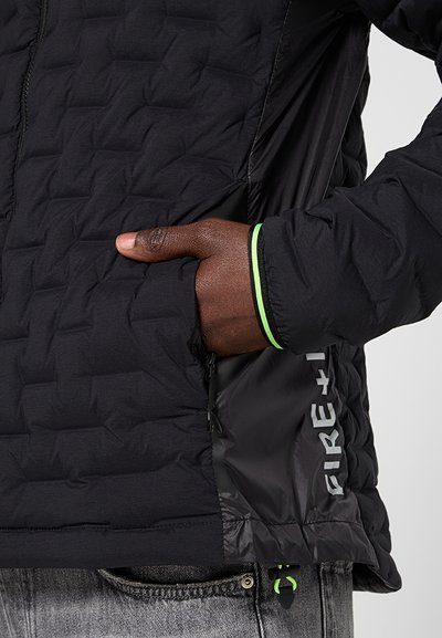 Sort quiltet jakke med tekstureret finish, en lynlås lomme og grønne detaljer ved manchetter og kant, med reflekterende branding på siden.